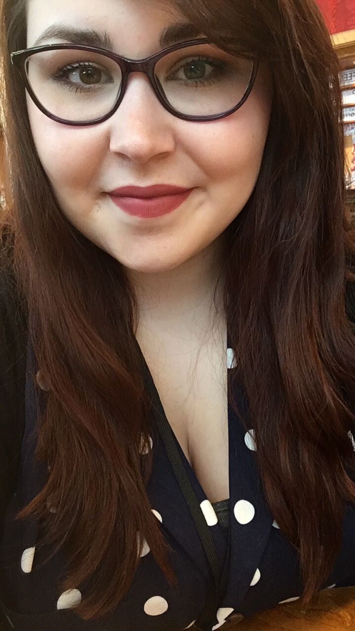 A selfie style image of Holly a 21 year old with long brown hair and purple slightly rounded glasses on. Holly is smiling at the camera and is wearing a nude/pink/brown lipstick. Holly is also wearing a dark blue shirt with medium white spots on it. There is also a black lanyard around her neck.