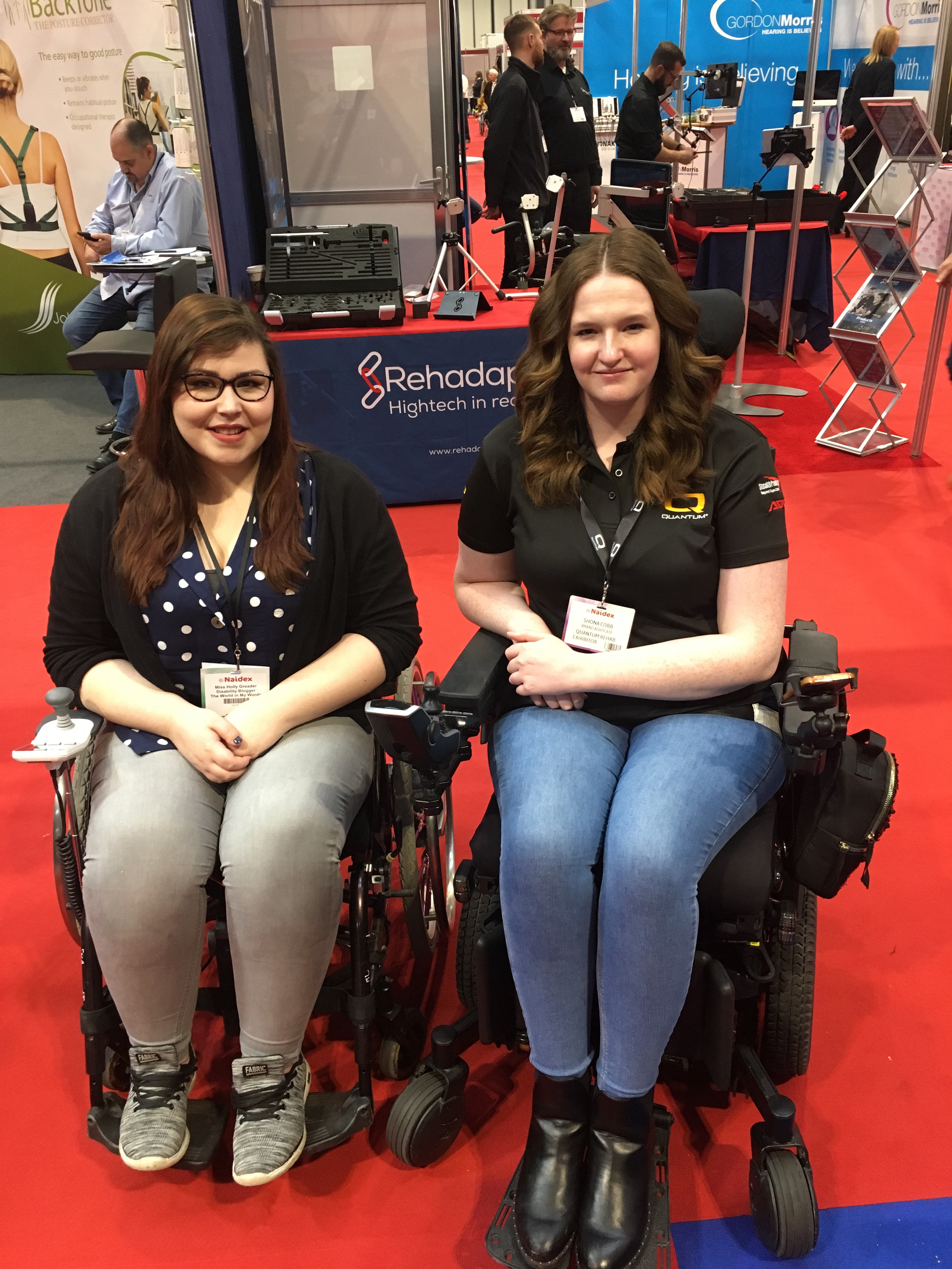 A imagine of two women in their wheelchairs on red carpet with stands behind them. On the left Holly sit in her wheelchair with her hands on her lap. Wearing a blue shirt with white Polka dots and a black Cardigan. A black lanyard is hanging around her neck with her name on it. On the right is Shona she is sitting in her wheelchair with her hands to the side. She is wearing light blue jeans and a black polo neck top. She also has a black lanyard around her neck with her name on