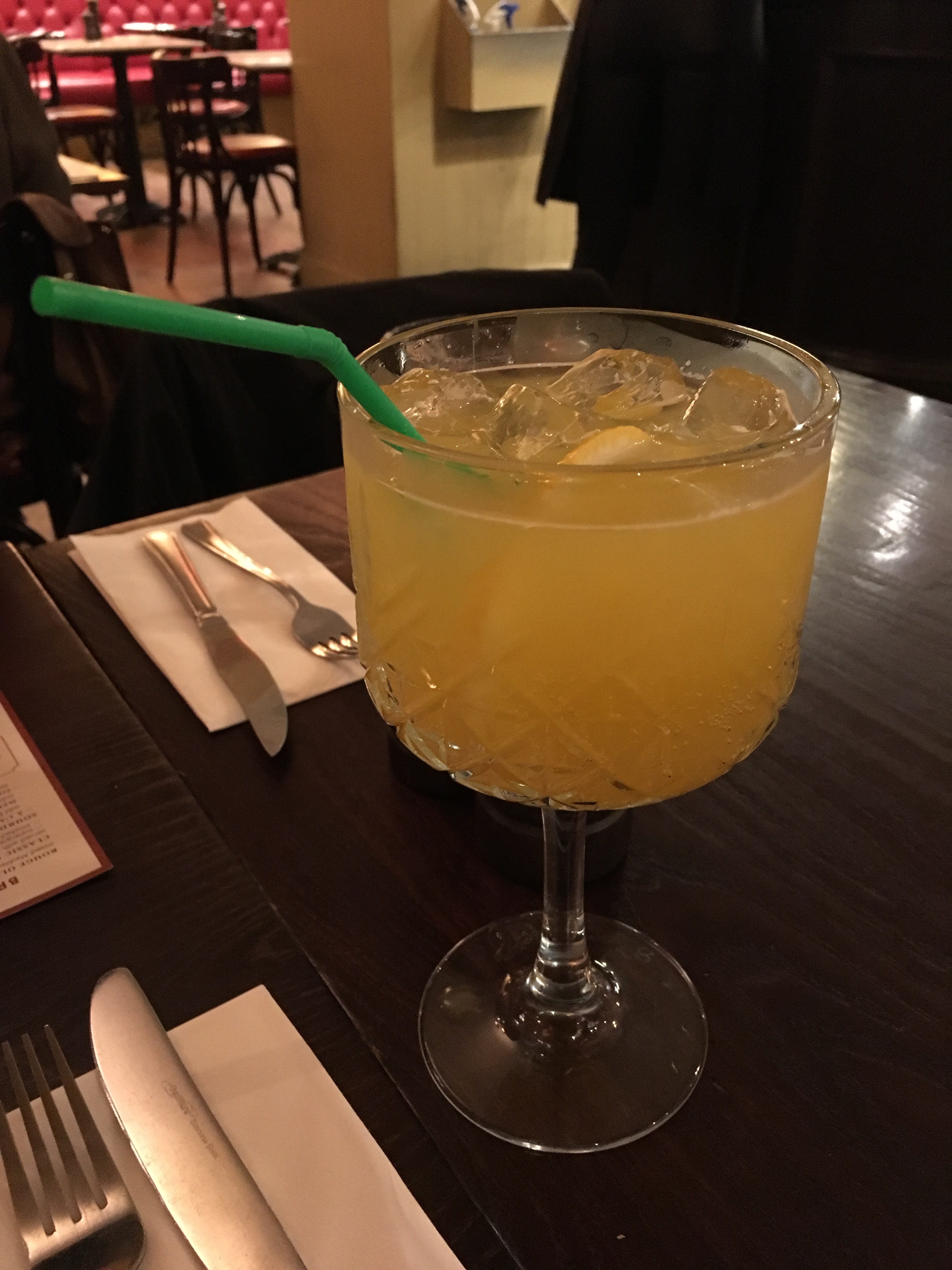 A brown table with two napkins, knives and forks. On the right is a tall large glass with a yellow coloured liquid and ice cubes inside. As well as green plastic bendy straw.