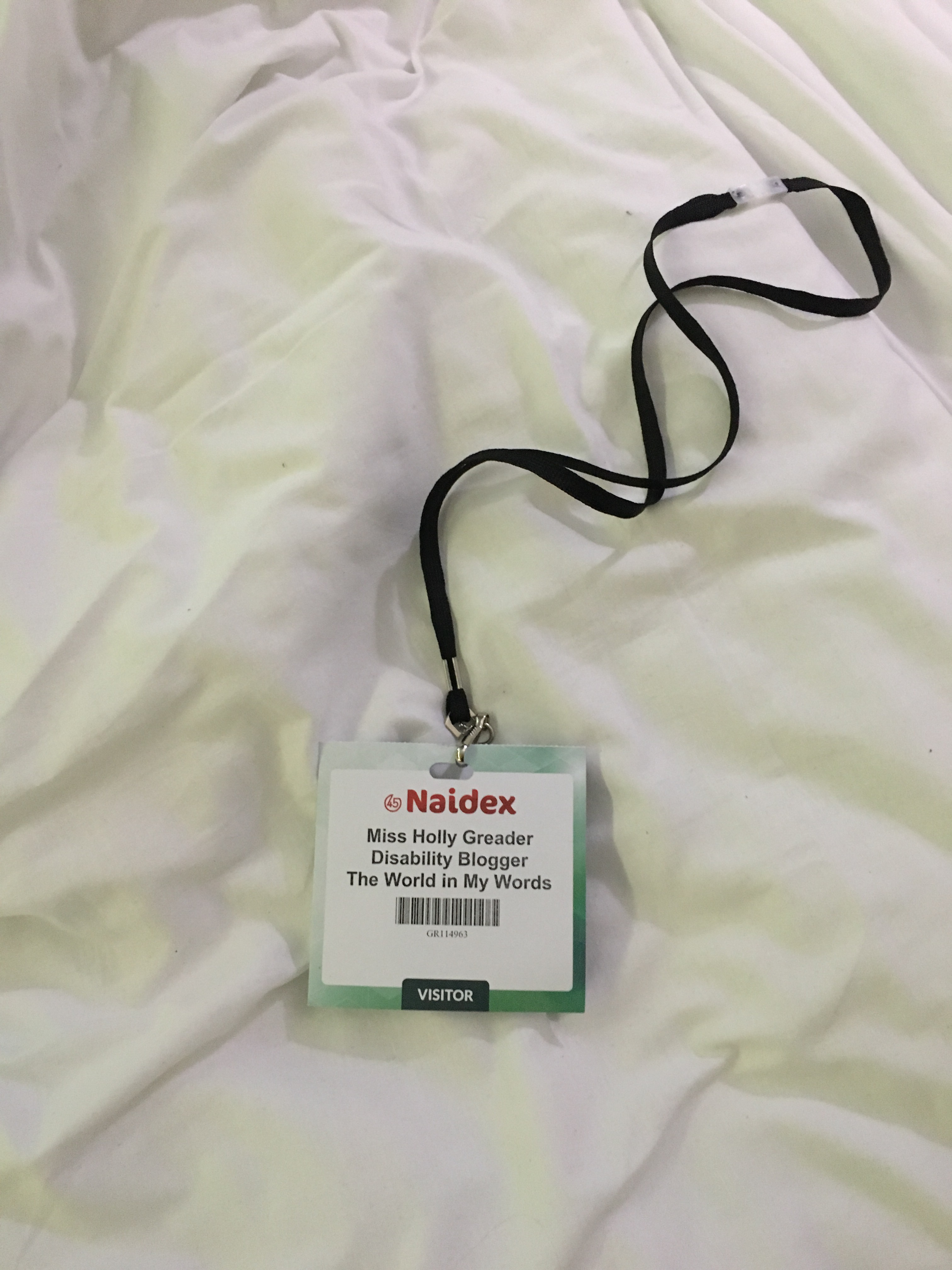 A pictures of a white bed sheet with a thin black lanyard on top with a piece of card attached, the a blue border. The card reads Naidex, Miss Holly Greader, Disability Blogger, The World in My Words. It also has a barcode underneath.