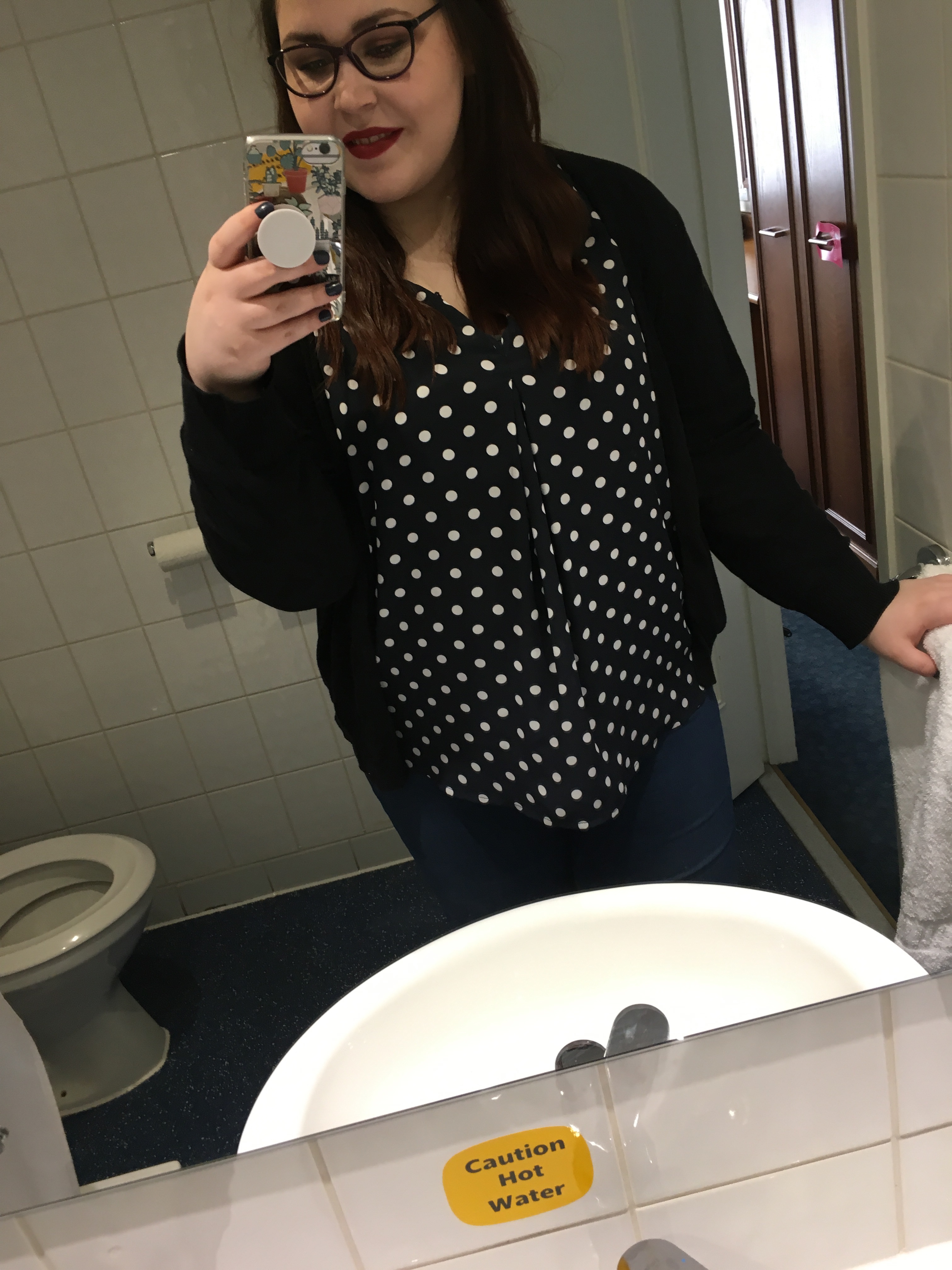 Holly is taking a selfie in a bathroom mirror. You can see part of a white sink and a toilet. Holly is holding onto a towel and towel rail on the right. She is wearing dark blue jeans with a dark blue blouse with white Polka dots and a black Cardigan. Her hair is long and brown she is also wearing red lipstick and purple rounded glasses. Her phone is also in her hand taking the picture.