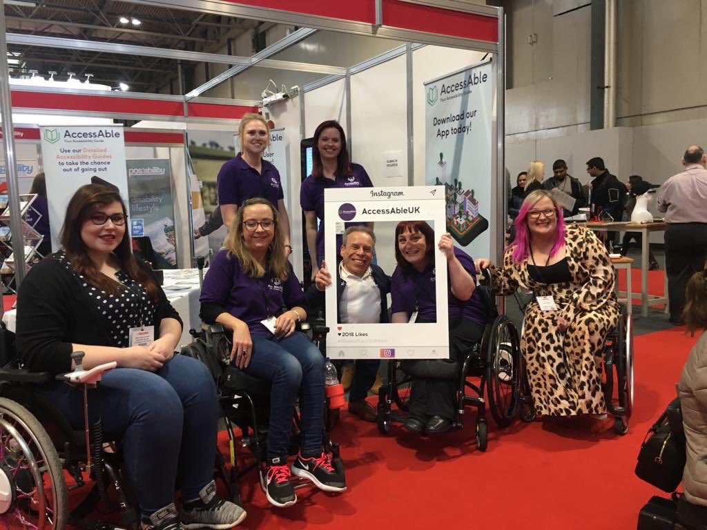 A group photo in front of the AccessAble stand. On the left are Holly and Carrie-Ann sat in their wheelchairs, in the middle is Warwick Daivs and Lucy in her wheelchair together holding an AccessAble Instagram cut out board. Behind them stands two of the AccessAble team. On the right sits Emily Yates. Everyone is smiling at the camera.
