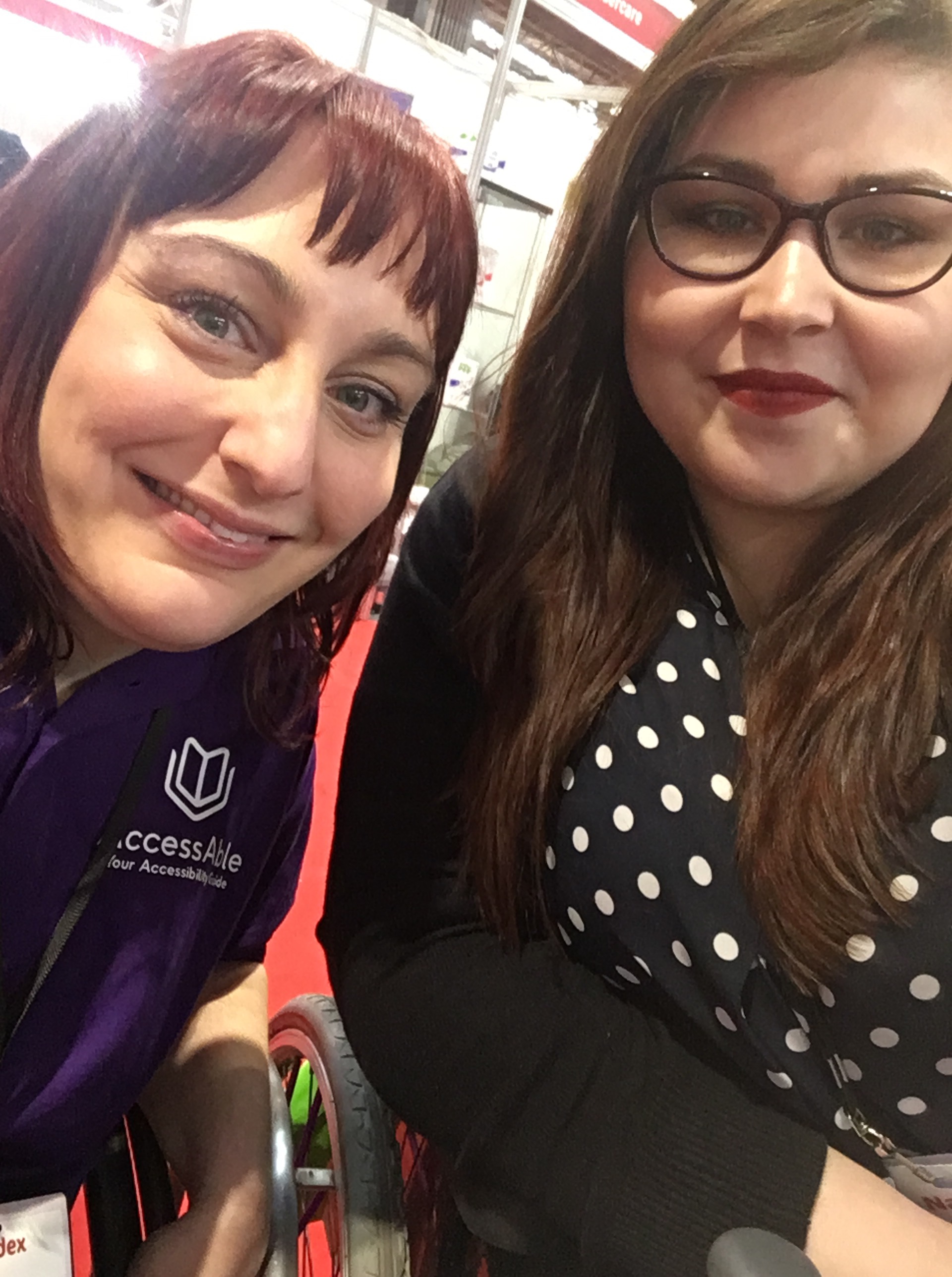 Two women sitting next to each other both leaning on the arms of their wheelchairs. On the left is Lucy with brown/red hair she is smiling at the camera and wearing a purple polo shirt which says AccessAble on it. On the right sits Holly she has long brown hair, slightly rounded purple glasses, she is wearing red lipstick. She is wearing a dark blue blouse with white polka dots and a black cardigan.