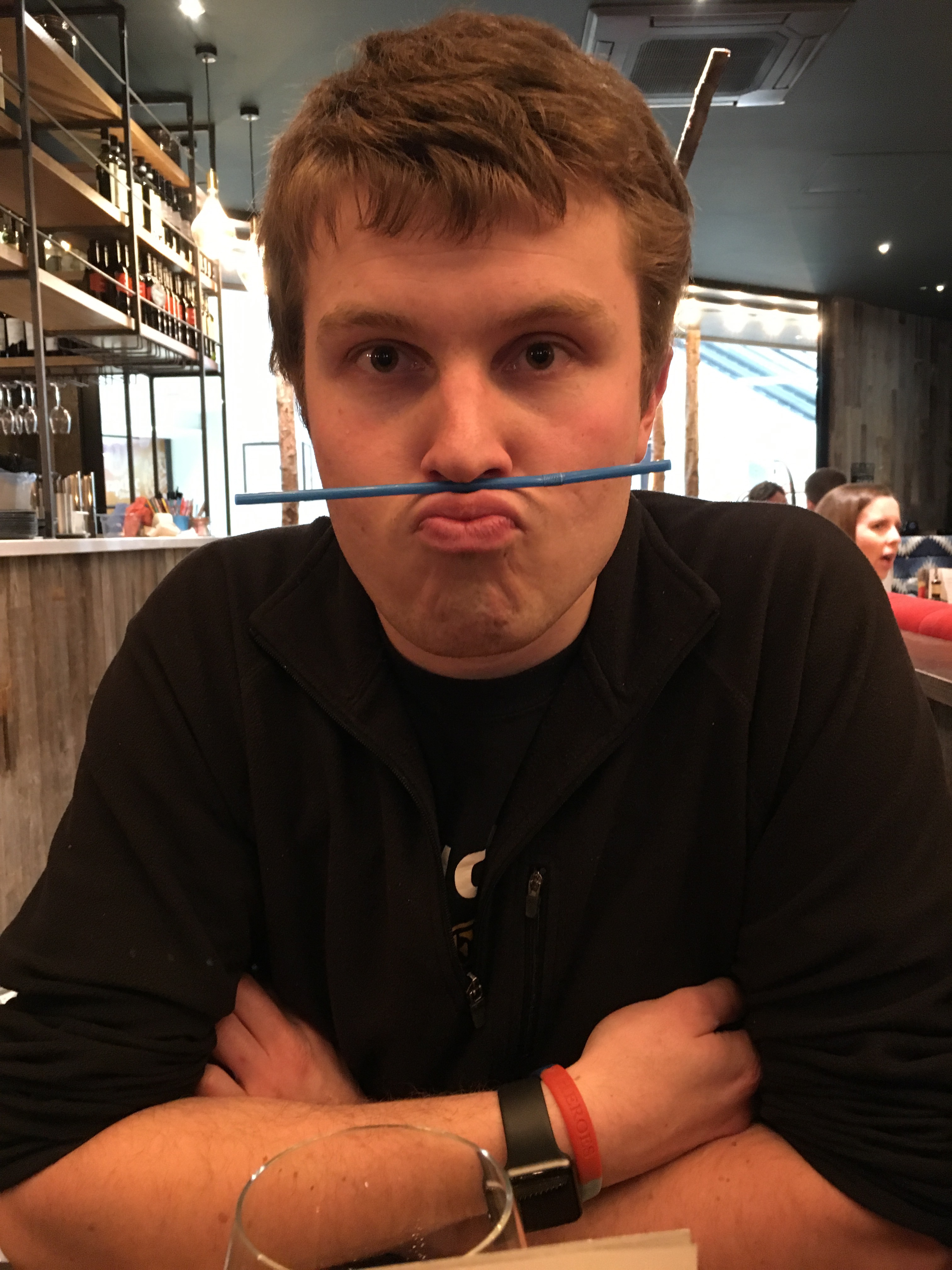 A photo of James a 23 year old with slightly wavy short brown hair. He has his arms crossed and is wearing a black jumper. He is pursing his lips pushing them out and up to hold a straw between his lips and nose. In the background you see other restaurant customers and part of the bar.