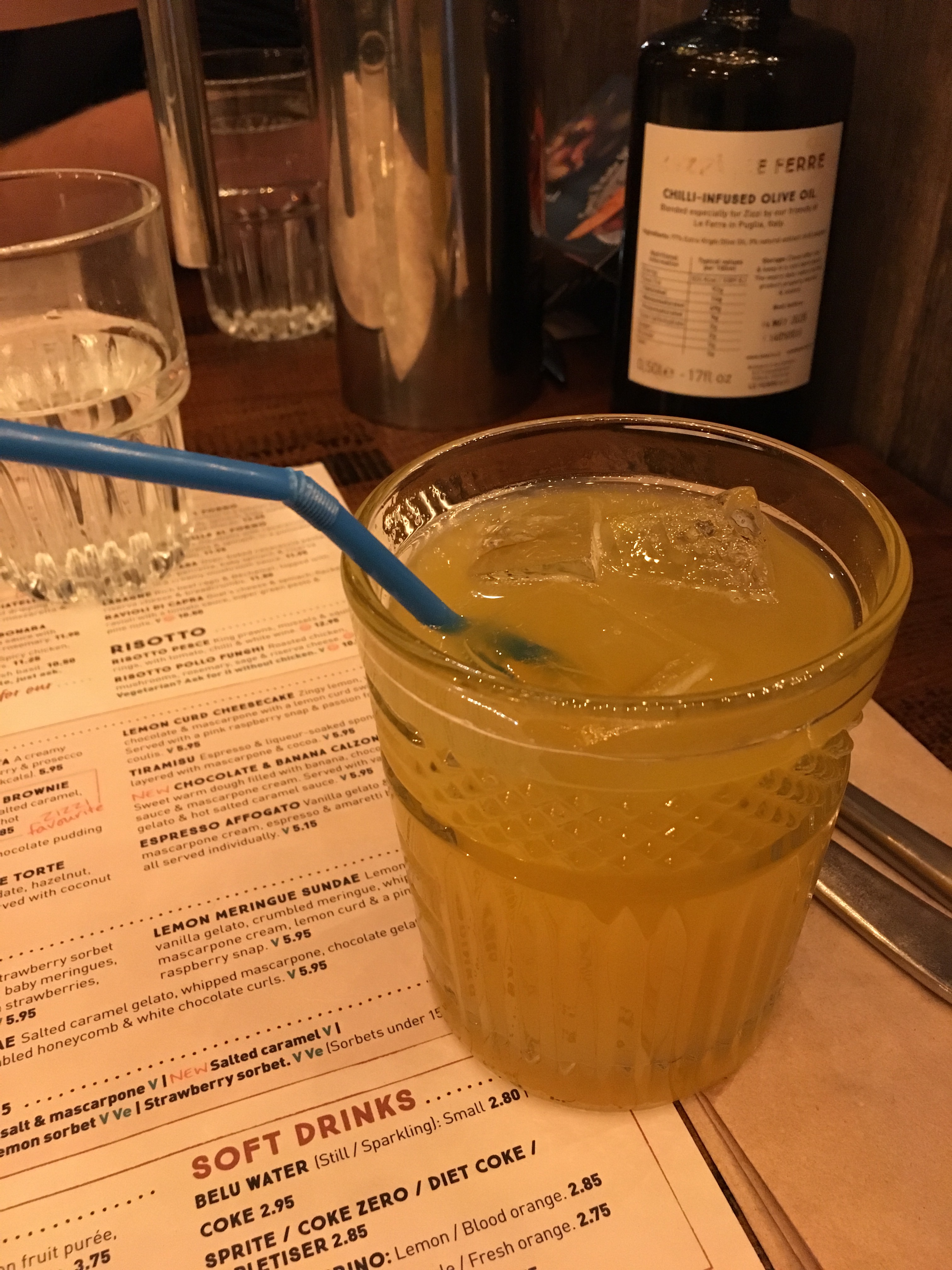 A short decorative glass with a yellow/orange liquid and ice in as well as a blue plastic bendy straw. The glass is sat on a menu and white napkin. There are other general restaurant item in the background including a water glass, olive oil and water jug.