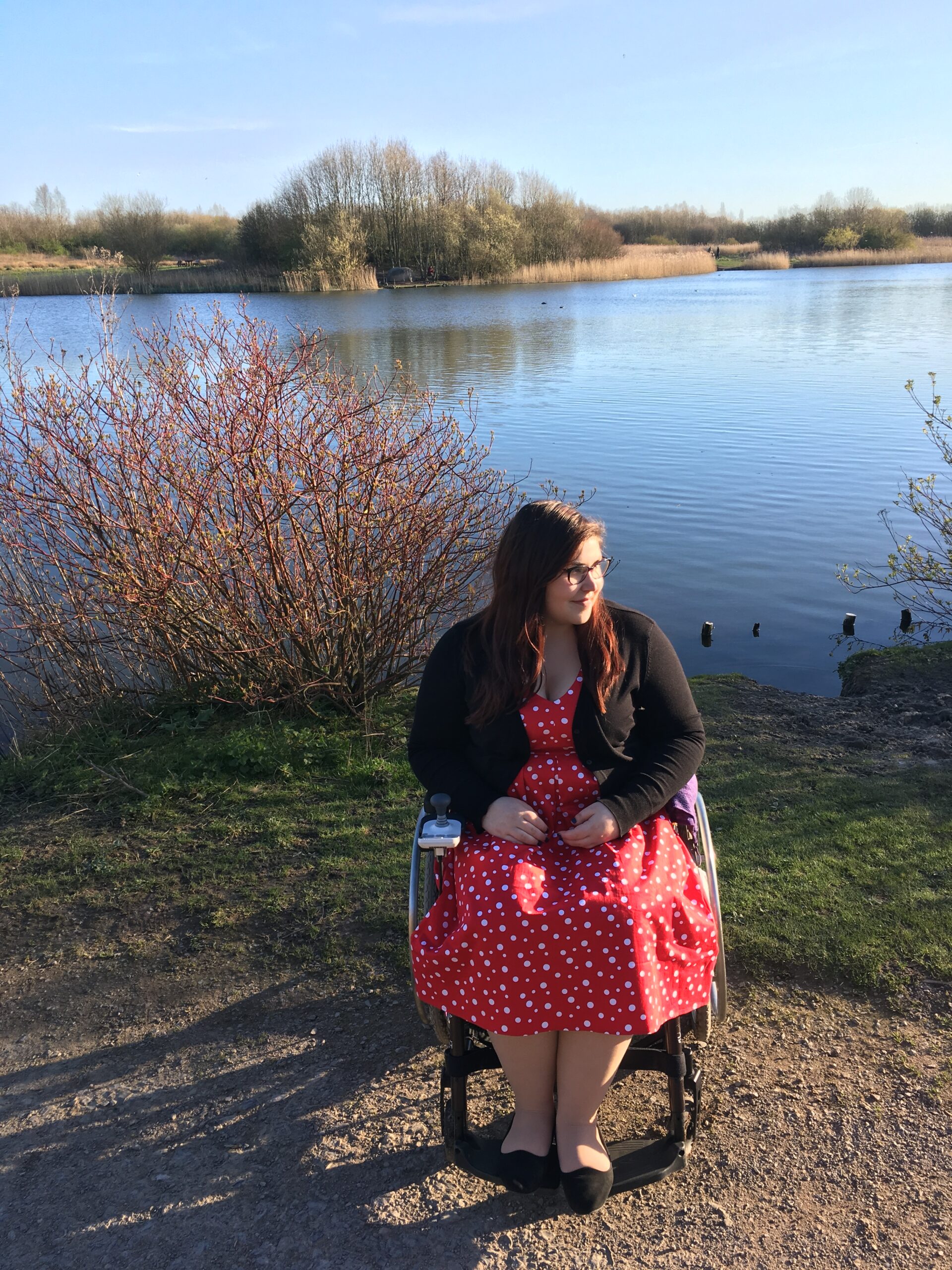 Holly is sitting in her wheelchair. She is wearing a red dress with a black cardigan. She's on a gravel path with a large blue lake behind her and green scenery.