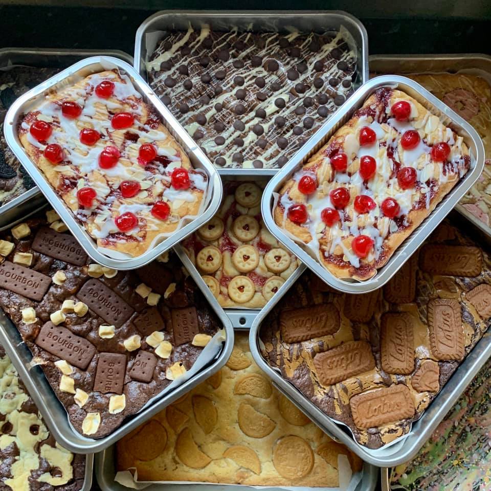 Eleven small baking trays stack decoratively around and on top of another. You can see blondies and brownies in each tray. Some decorated with cherries and icing. Some chocolate with chocolate chips. Others with lotus biscuits, bourdon biscuits, party rings or oreos on top.  