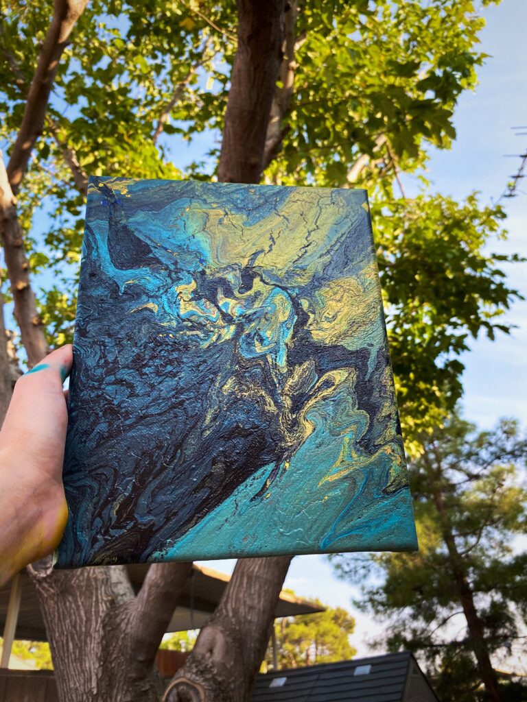 A canvas of abstract flow arts with blue, black, gold colours is being held up so you can see the blue sky and trees behind it. 