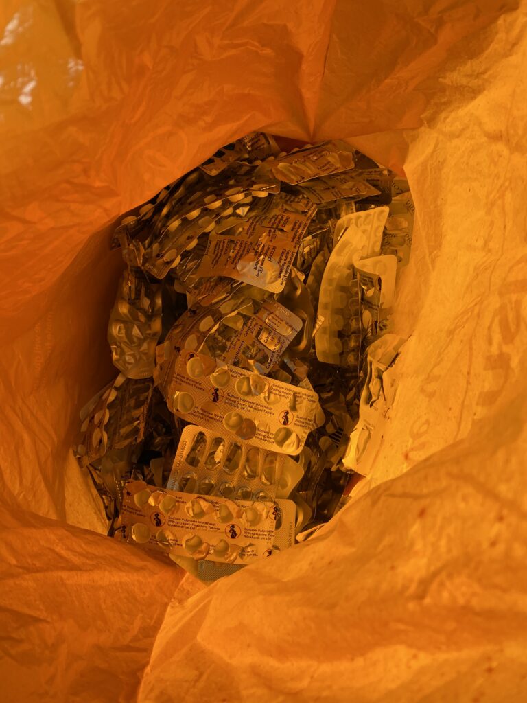 Inside a orange carrier bag is a big pile of empty medication packets. Each packet has a silver foil on the front and white plastic underneath. There is a slight hue to them from the bright orange bag.