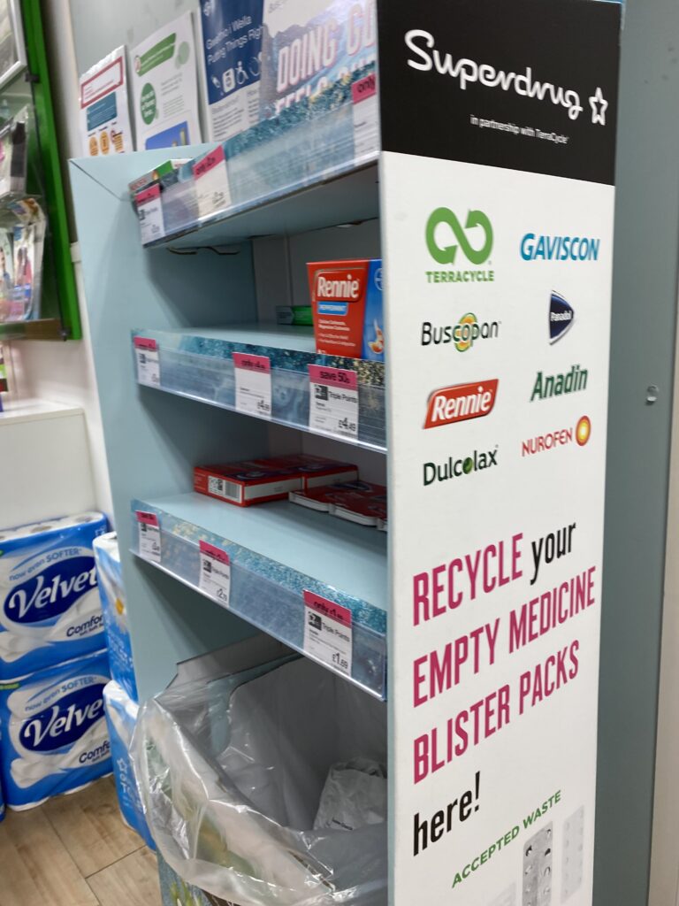 A cardboard shelving stand for products. On the side it says Superdrug with brands of medications below. There are medictions on sale on the stand as well as a bin for old medication packets.