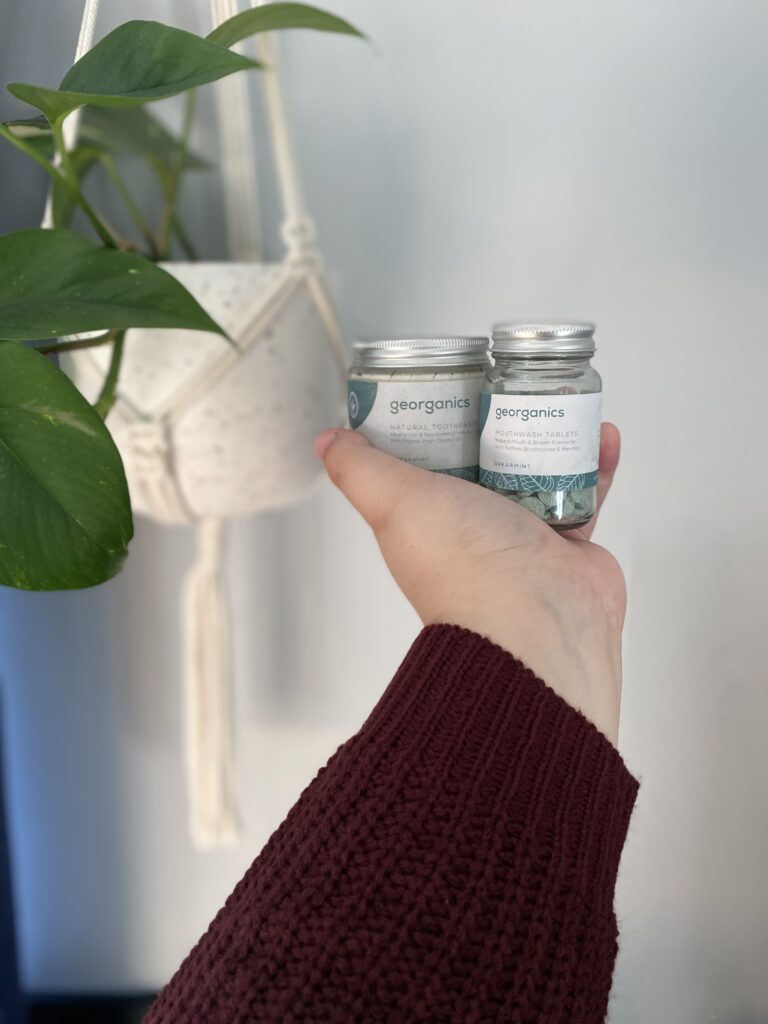 Grey wall in the background with a macrame plant hanger and a green plant flowing out. Georganics toothpaste jar and mouthwash tabs are being held in a hand where an arm is outstretched wearing a dark purple knitted jumper. 