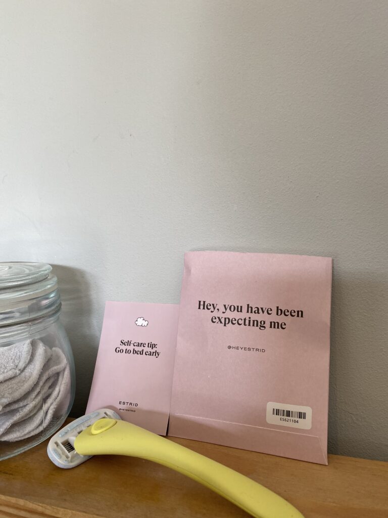 Grey background with a wooden ledge. On the ledge is a yellow steel razor behind is a pink packaging and an affirmation card. There is a glass make up pad jar on the left. 