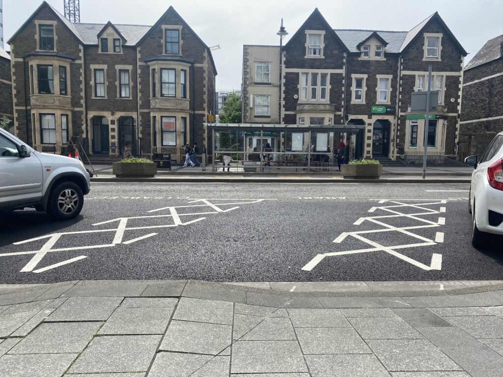 Buildings in the background and bus stops. There are diagonal blue badge parking bays. There are hatched areas on each side of the bay. The curb is high behind the bay with drop curbs on either side.