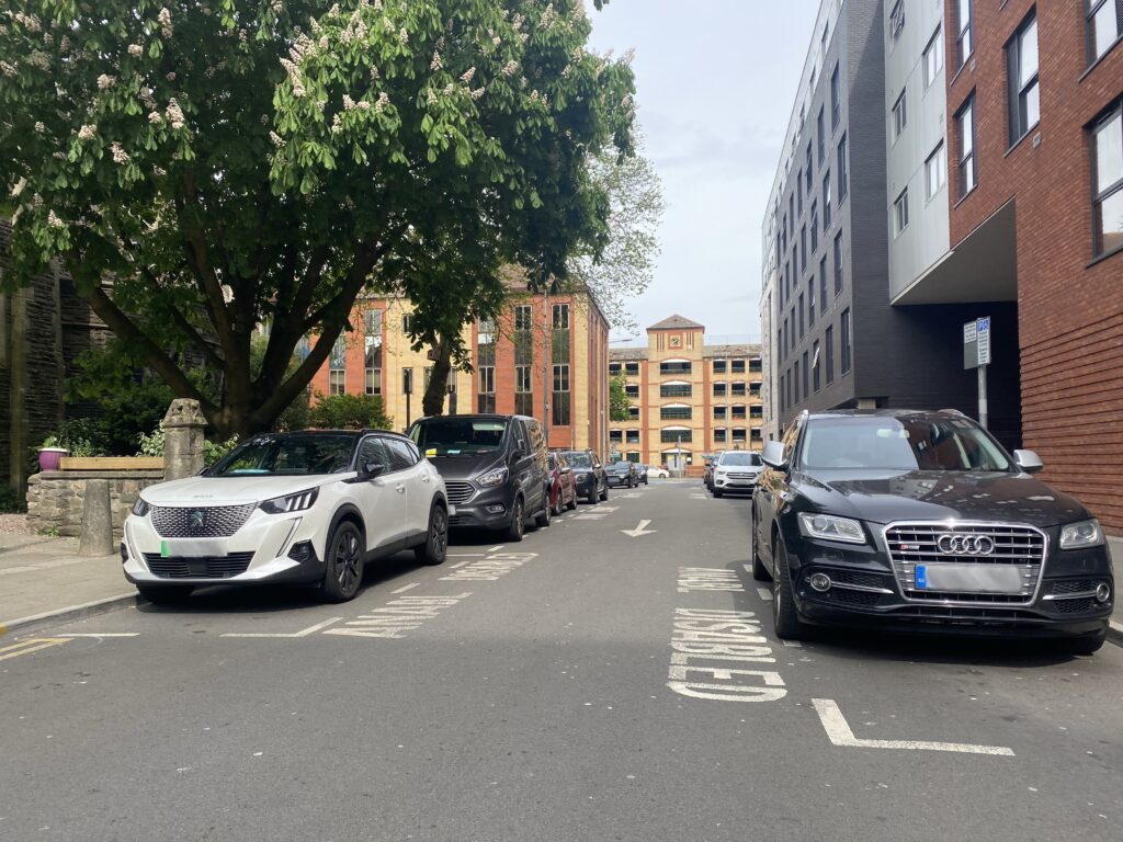 Buildings on both sides and at the end of the road. There are cars parked parallel down the road in disabled parking spaces also labelled in Welsh.