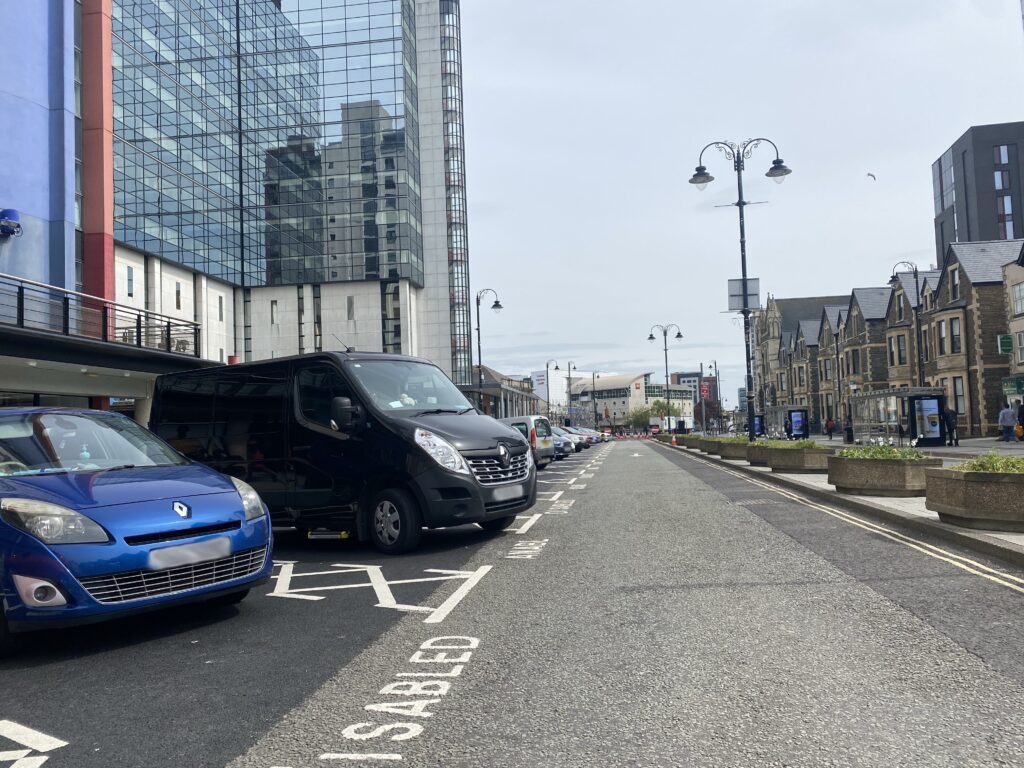 A long road with buildings on either side. To the left are accessible parking spaces labelled disabled in English and Welsh. There are cars parked all the down the road in the designated spaces.